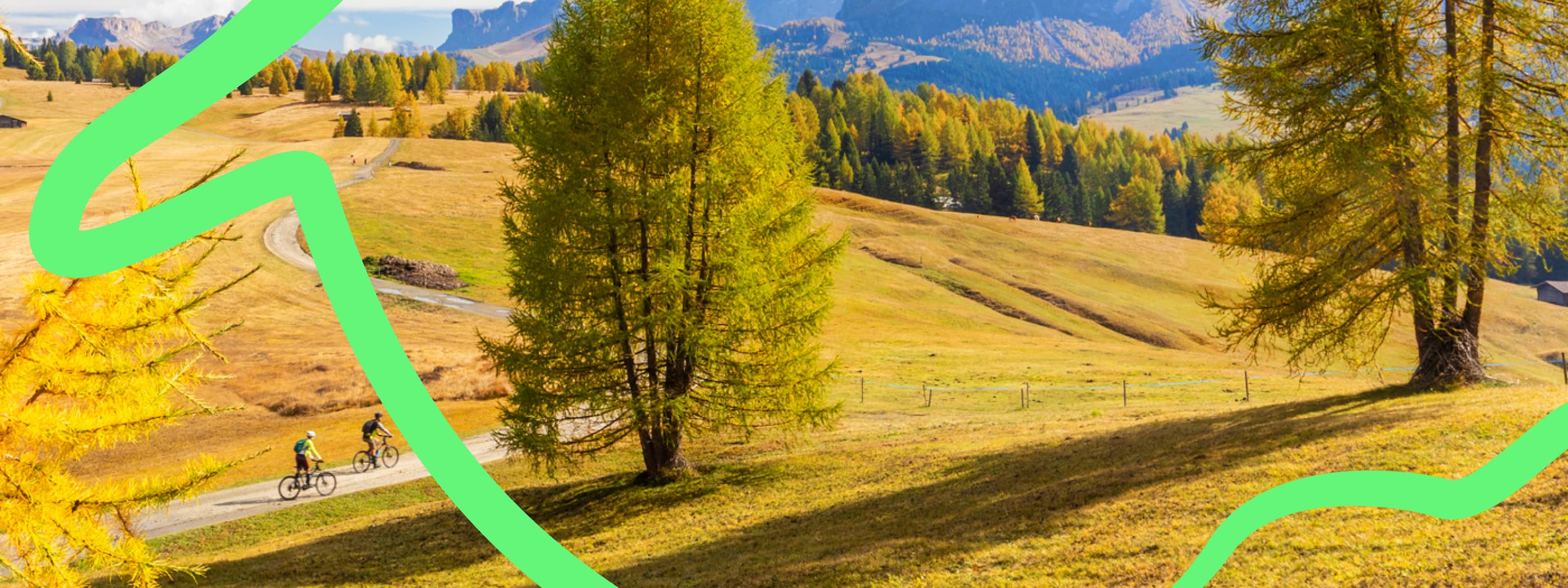 Two people ride bicycles along a dirt road winding between yellow grassy fields with small historic barns in front of a line of trees with colorful fall leaves and majestic bare rocky mesas.